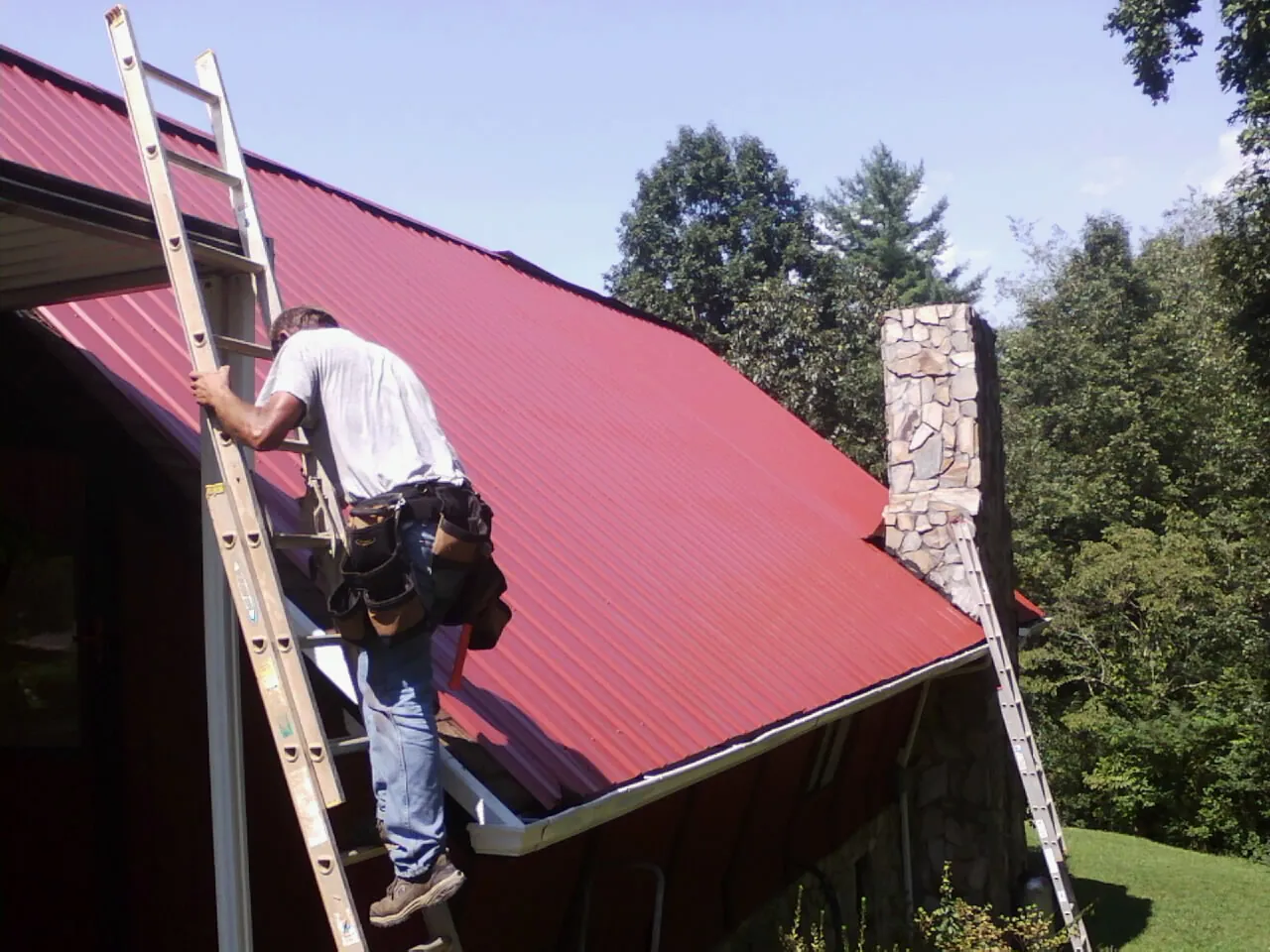 Roofer on a ladder beside a red metal roof and stone chimney — Soffit & Fascia Repair by Roofing Brothers in Beaver Dams